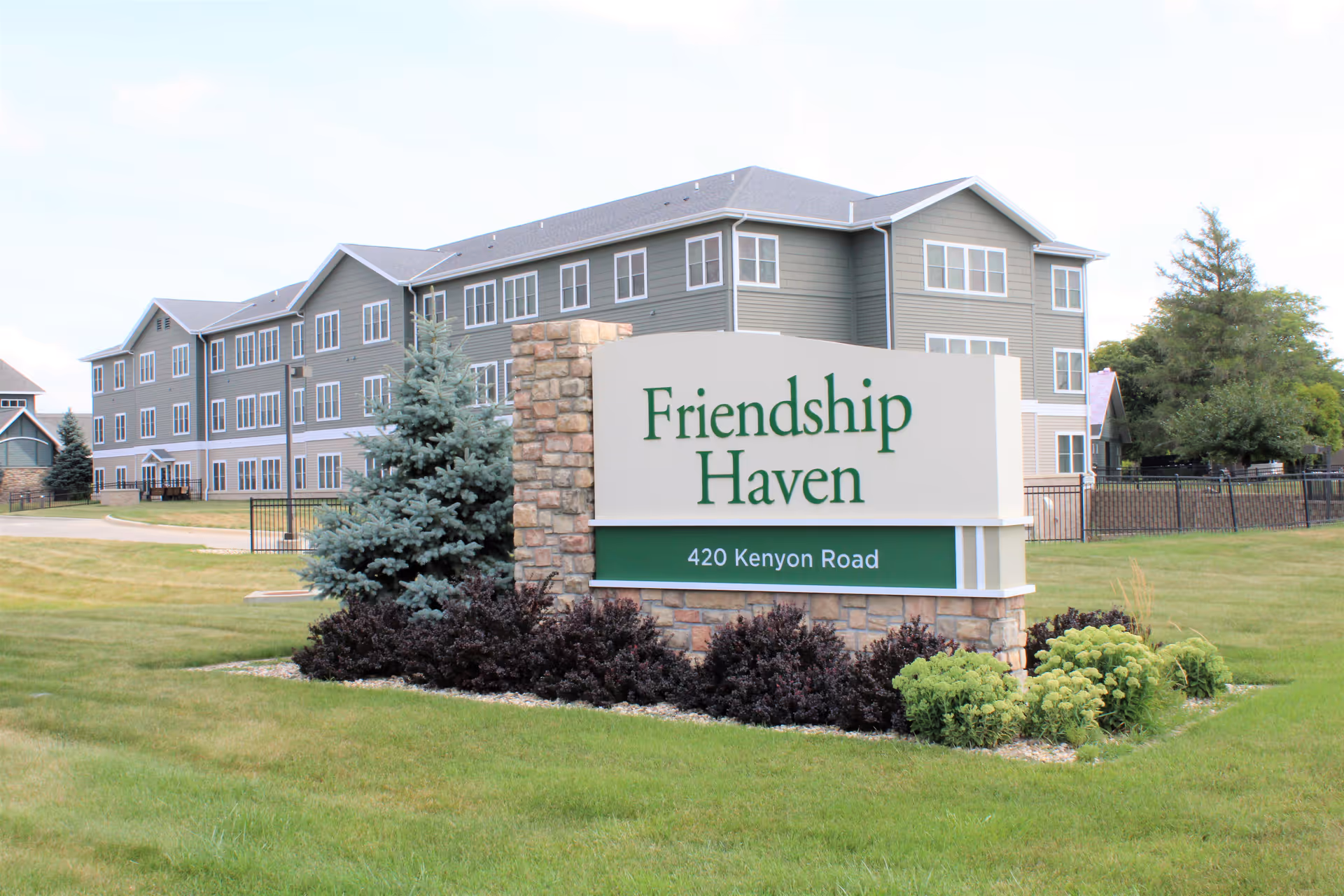 Exterior view of a multi-story senior living facility building with gray siding and many windows. In the foreground, there is a large stone and white sign with green text that reads 'Friendship Haven' and '420 Kenyon Road'. The area around the sign is landscaped with bushes and small trees, and there is a grassy lawn extending around the building.