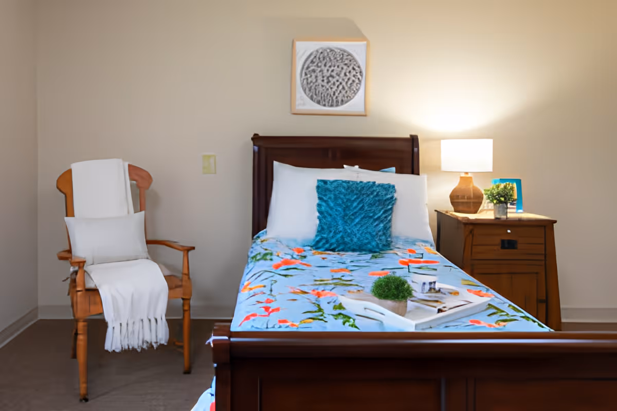 Single-bed bedroom with floral bedding, a wooden nightstand with a lamp, and a chair draped with a white throw.