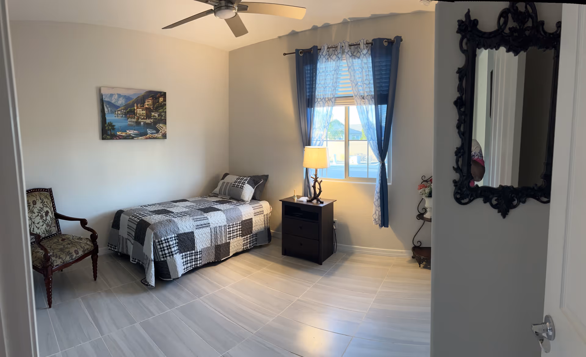 Well-lit single bedroom with a twin bed, nightstand and lamp, chair, window with curtains and a ceiling fan.