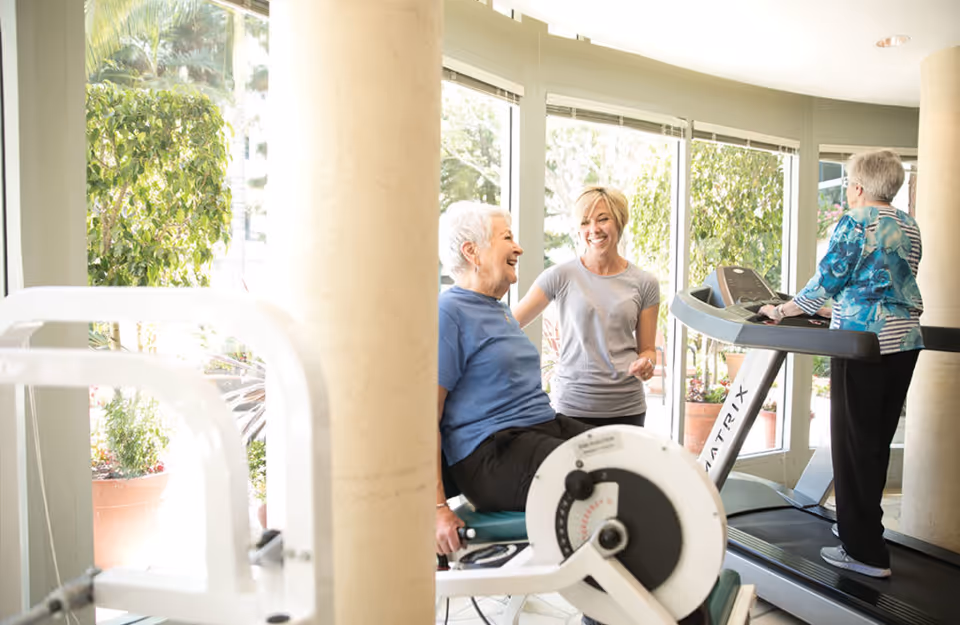 Two elderly women exercising in a fitness room with large windows showing greenery outside. One woman is using a seated exercise machine while smiling and talking with a younger woman standing beside her. Another elderly woman is walking on a treadmill in the background.