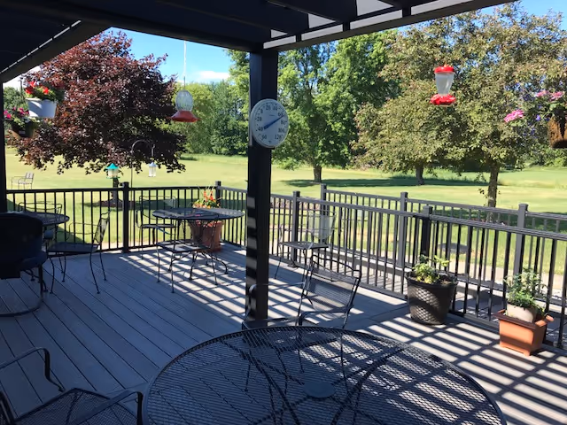 A shaded outdoor patio area with black metal tables and chairs, potted plants, hanging bird feeders, and a large thermometer mounted on a post. Beyond the patio is a grassy area with trees and a clear blue sky.