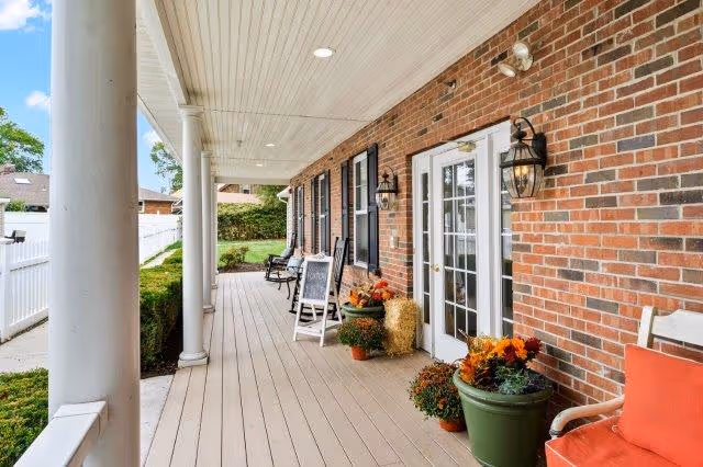 Covered brick front porch with white columns, chairs, potted plants and double glass doors.