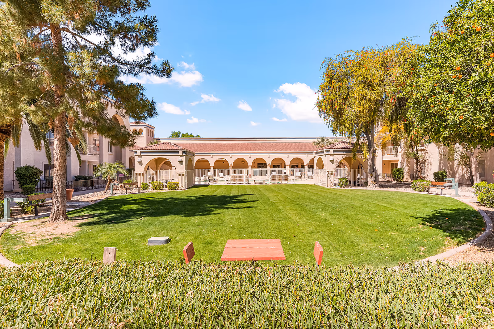 A green lawn courtyard with trees and a Mediterranean-style senior living building featuring an arched colonnade in the background.