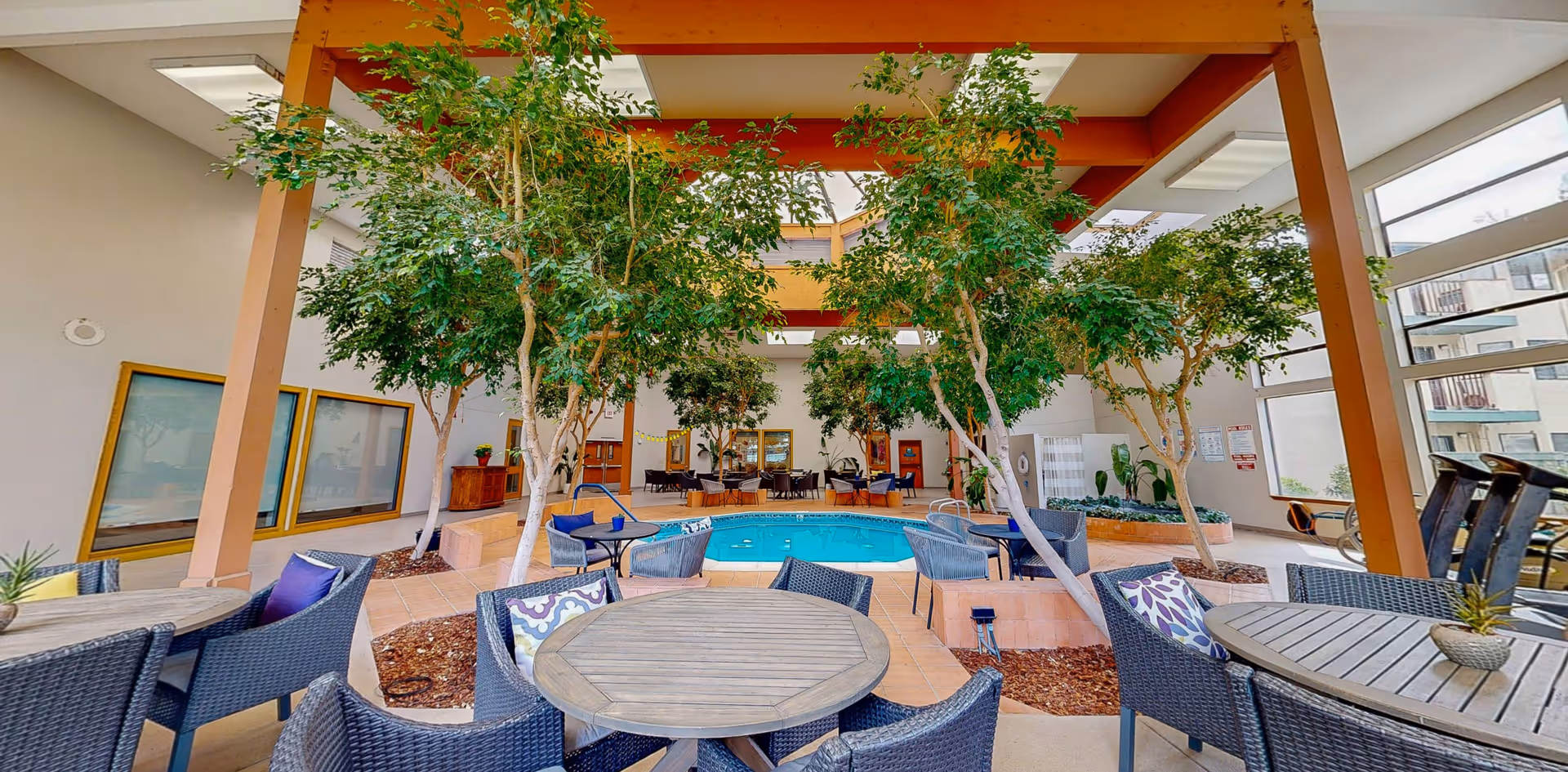 Indoor seating area with round tables and wicker chairs arranged around a small pool. The space is decorated with several tall indoor trees and has large windows allowing natural light to enter. The ceiling features skylights and wooden beams.