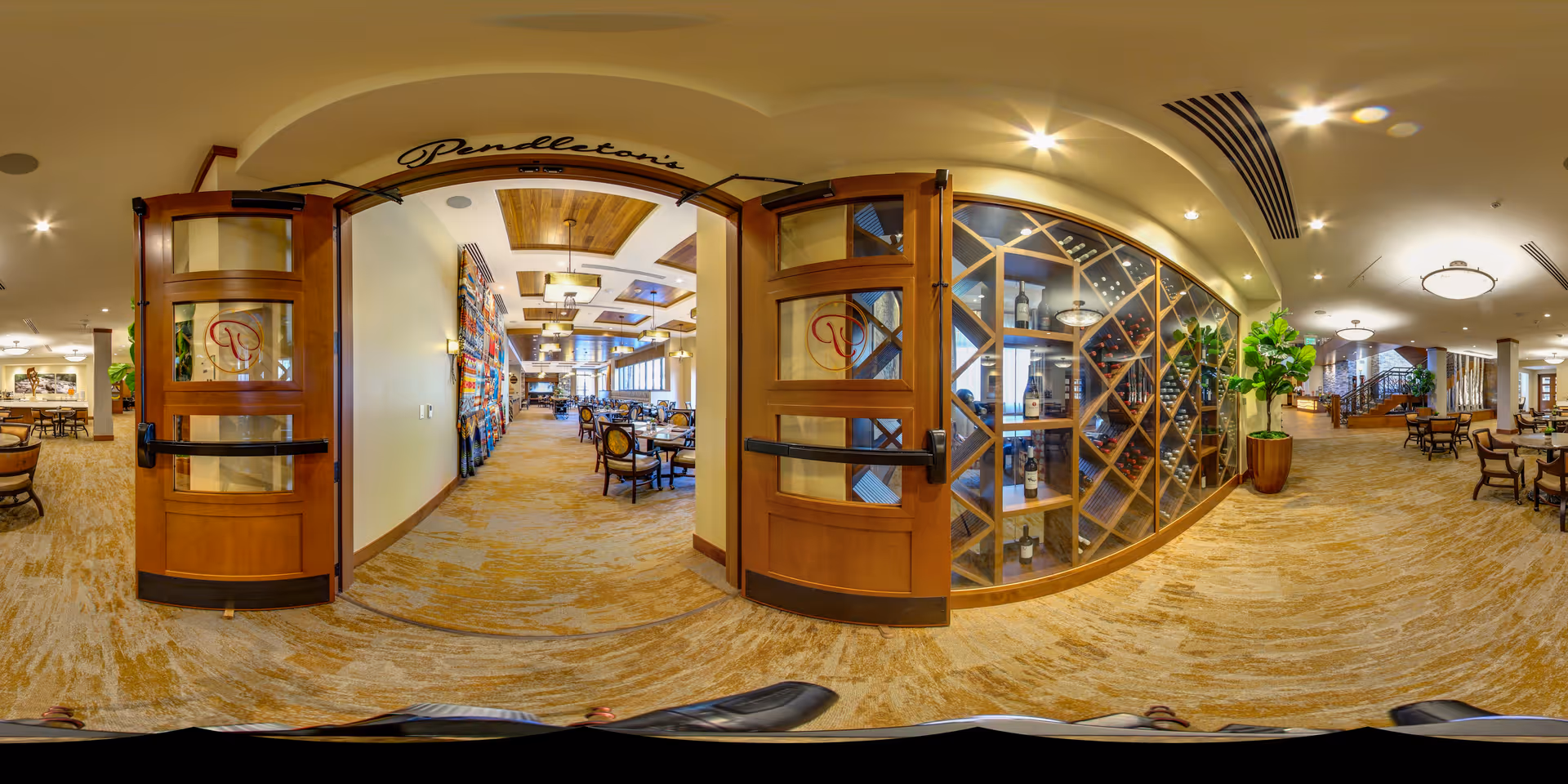 A spacious and well-lit dining area in a senior living facility with wooden double doors labeled 'Pendleton's' opening into a room with multiple tables and chairs. To the right, there is a large wine rack filled with bottles and decorative items. The carpet is beige with a subtle pattern, and the ceiling features recessed lighting and wooden accents. There are plants and additional seating areas visible in the background.