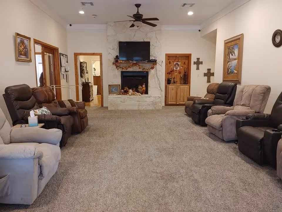 A cozy living room with a carpeted floor, featuring two rows of recliner chairs facing each other. At the far end, there is a stone fireplace with a flat-screen TV mounted above it. The walls are decorated with framed paintings and crosses, and there are two wooden doors and a cabinet near the fireplace.