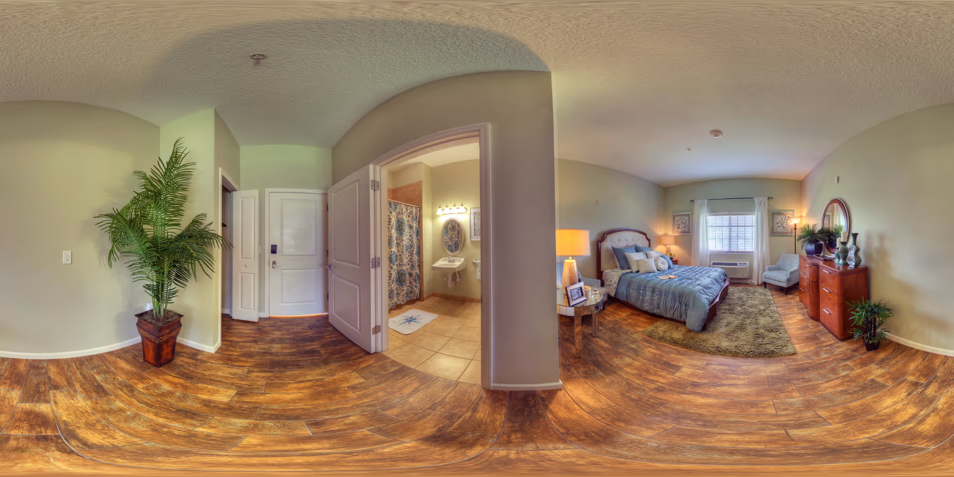 A panoramic view of a senior living apartment interior showing a bedroom and a bathroom. The bedroom features a bed with blue bedding, a nightstand with a lamp and photo frame, a dresser with decorative items, a chair, and a window with white curtains. The bathroom has a tiled floor, a shower with a patterned curtain, a sink with a mirror above it, and a bath mat. The flooring throughout is wood-style, and there is a large potted plant near the entrance door.