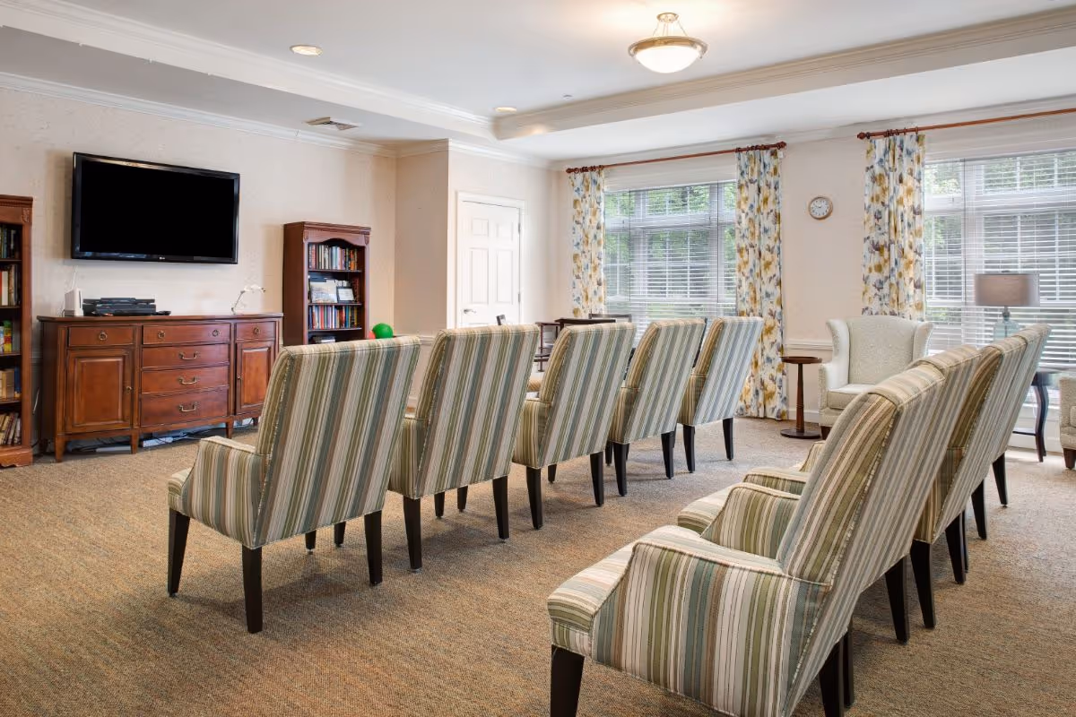 A well-lit common room with two rows of striped upholstered chairs facing a wall-mounted flat-screen TV. The room has large windows with floral curtains, a wooden cabinet, bookshelves, and additional armchairs near the windows.