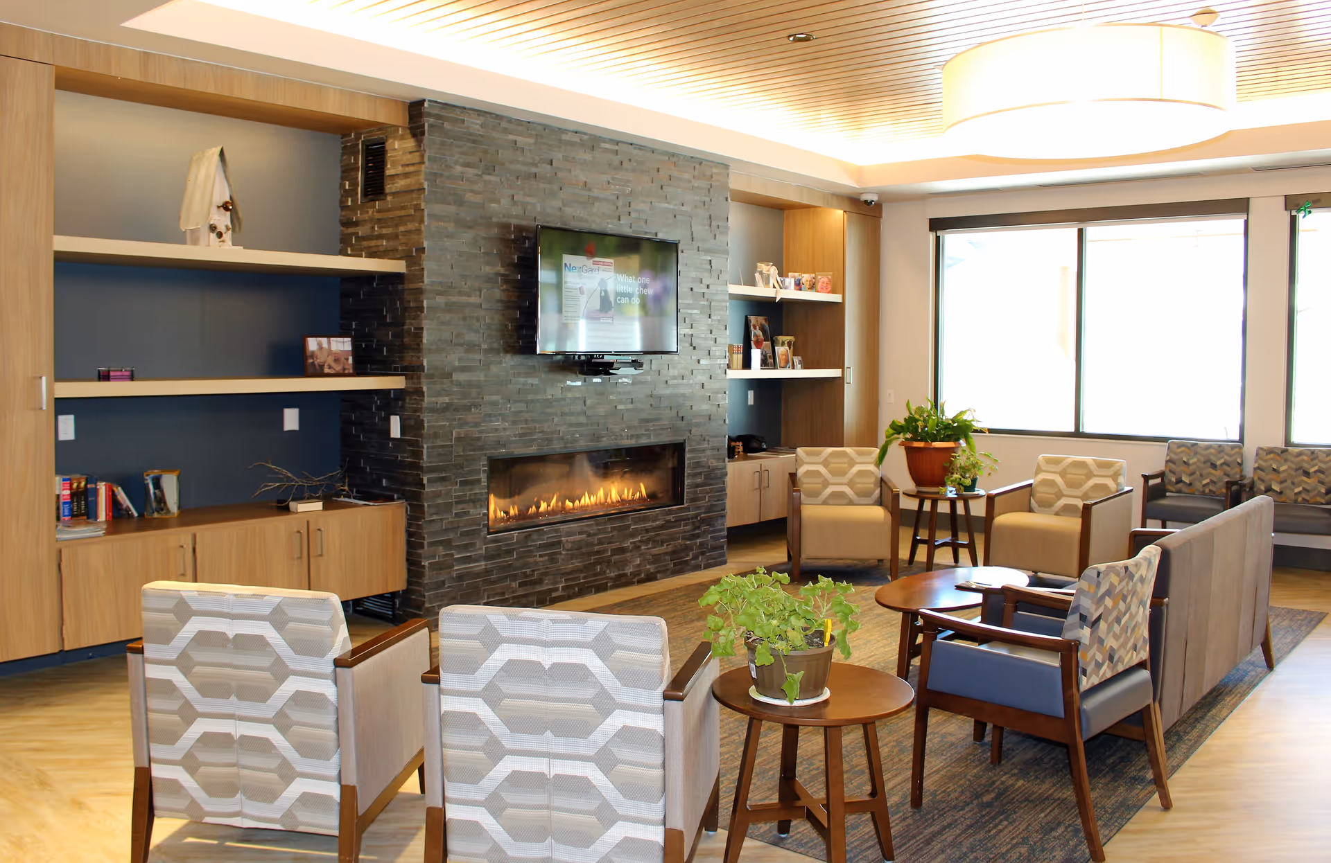 Bright senior living common lounge with a stone fireplace, wall-mounted TV, multiple armchairs and side tables, and large windows.