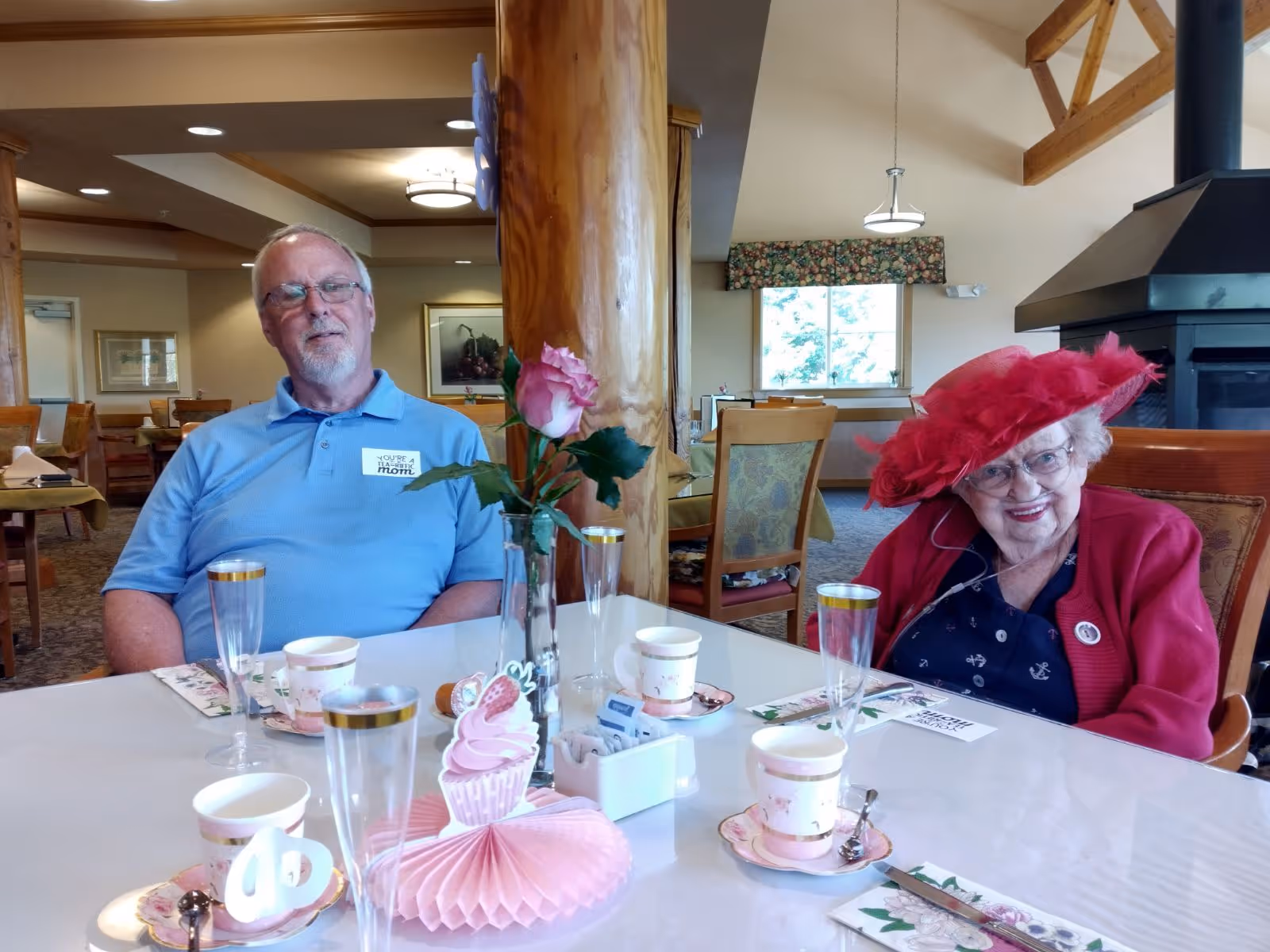 An elderly woman wearing a bright red hat and red cardigan sits at a dining table with a man in a light blue polo shirt. The table is set with teacups, saucers, and decorative items including a pink paper cupcake decoration and a vase with a pink rose. The background shows a spacious dining area with wooden furniture and a large window.