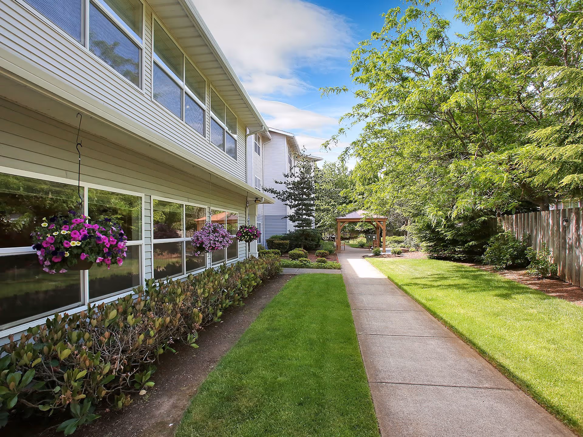 Outdoor walkway alongside a senior living facility building with hanging flower baskets, green bushes, a well-maintained lawn, trees, and a wooden gazebo in the distance under a partly cloudy blue sky.