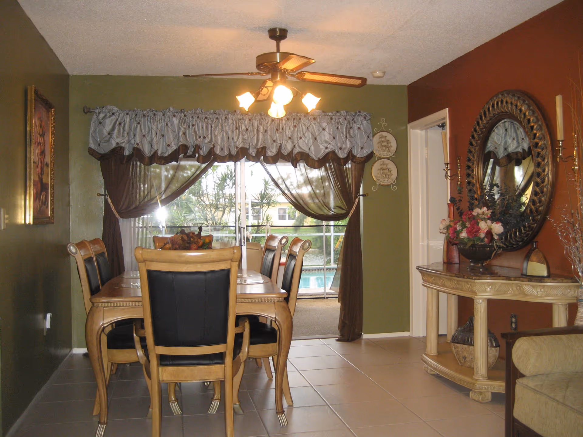 Dining room with a wooden table and six chairs with black cushions. A ceiling fan with lights is above the table. The room has green and brown walls, with a large window covered by curtains showing an outdoor pool area. A decorative console table with a round mirror and flower arrangement is on the right side.