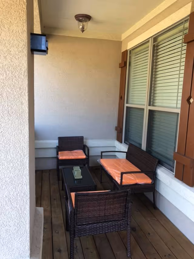 A small covered outdoor patio area with wooden flooring, featuring a set of dark wicker furniture including two chairs, a loveseat, and a glass-top coffee table. The chairs and loveseat have orange seat cushions. The patio is adjacent to a wall with a window that has closed blinds and decorative wooden shutters. A ceiling light fixture is mounted above.