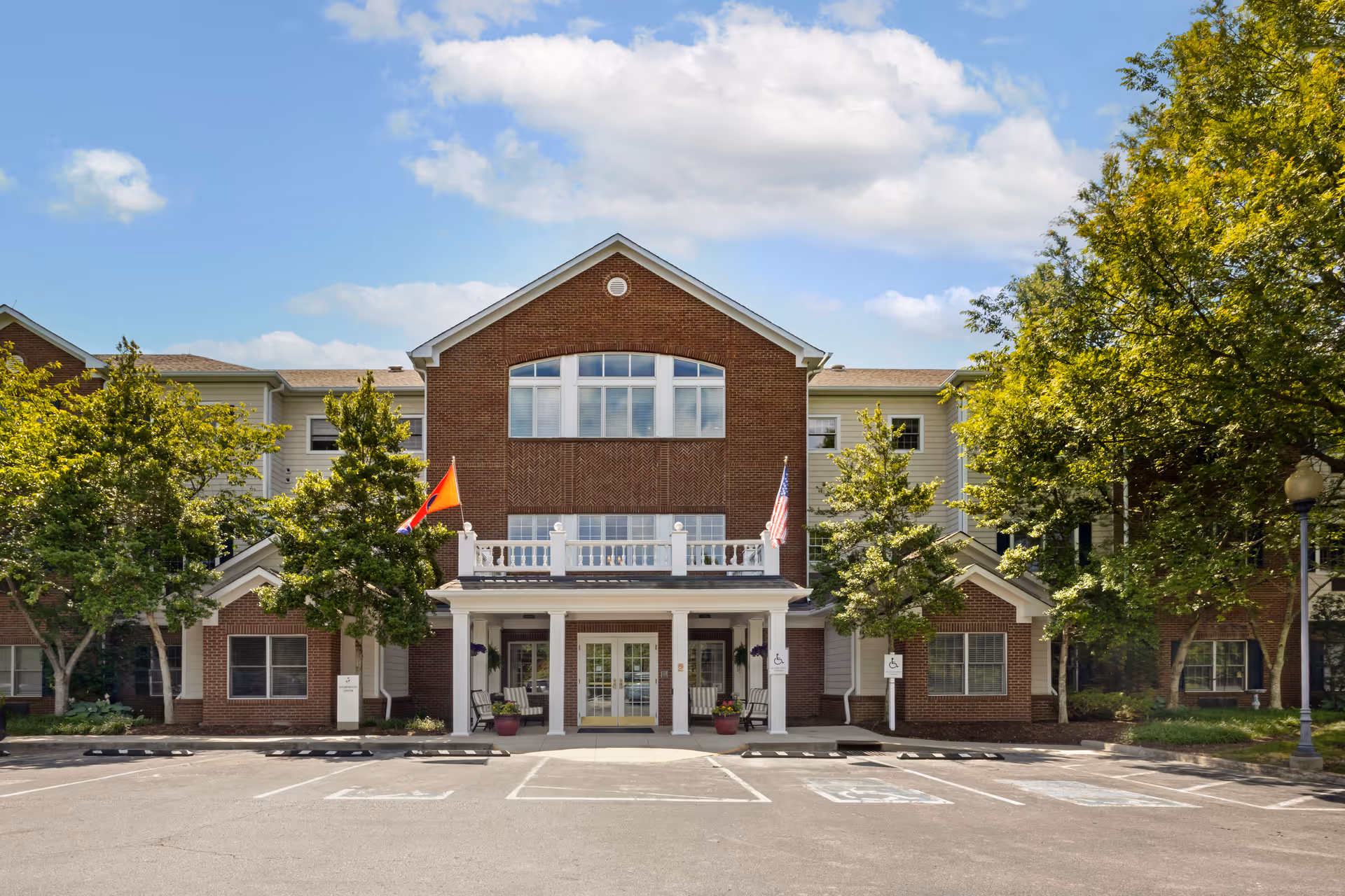 Front exterior view of a senior living facility building with a brick and beige facade, two flags on poles near the entrance, several trees on either side, and a parking lot with handicap spaces in front.