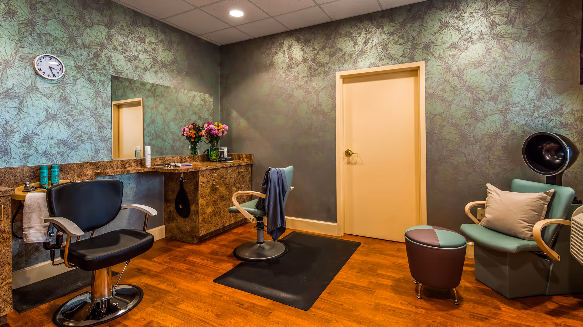 Interior view of a salon room with two styling chairs, a large mirror, a countertop with hair products and flowers, a clock on the wall, and a seating area with a chair, ottoman, and a hair dryer hood. The walls have a green leafy patterned wallpaper and the floor is wooden.