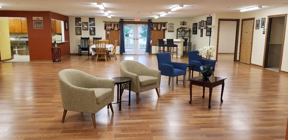 A spacious room with wooden flooring featuring two beige armchairs and two blue armchairs arranged around small tables with a flower vase. In the background, there is a dining area with wooden chairs and tables, a kitchen area to the left, and double glass doors with curtains leading outside. The walls are decorated with framed pictures and there are several doorways visible.