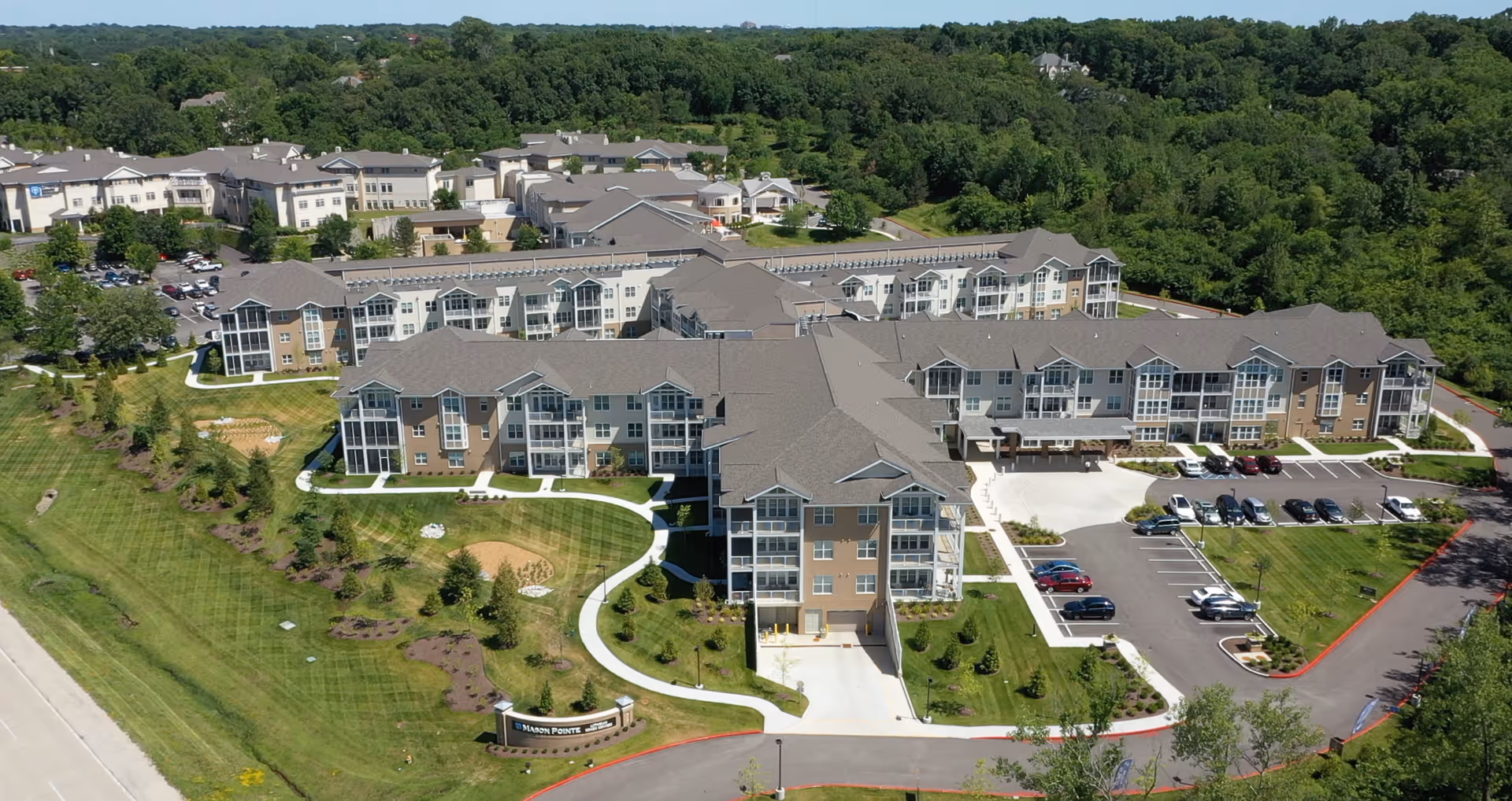 Aerial view of a large multi-wing senior living facility with landscaped lawns, walking paths, and a parking lot surrounded by trees.