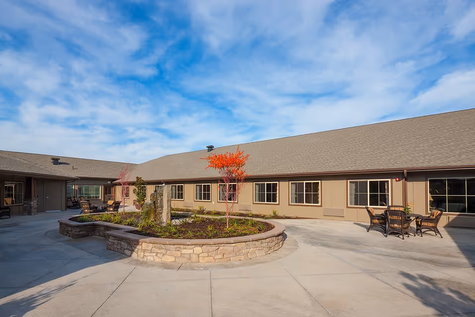 Sunny courtyard of a single-story memory care building with a circular raised stone planter, outdoor seating, and a clear blue sky.