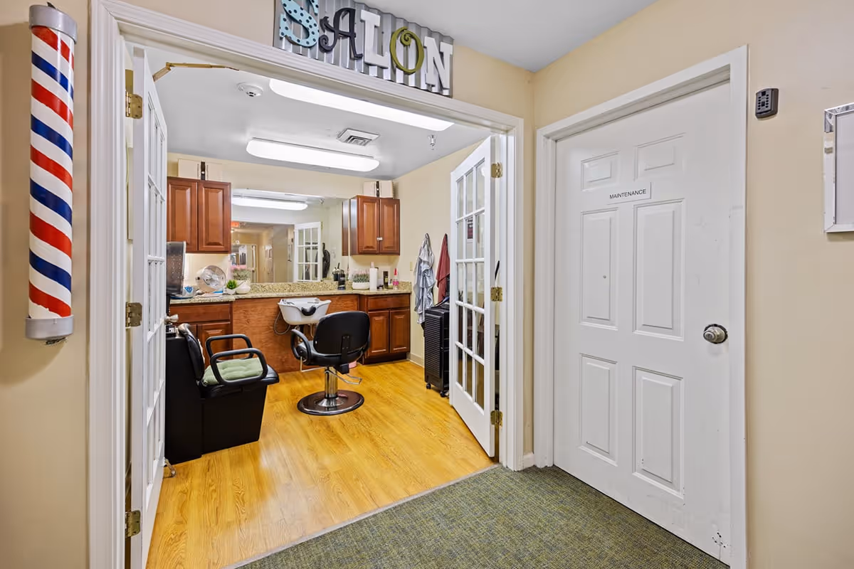 Interior view of a salon room in a senior living facility with wooden flooring, a black salon chair, a shampoo station, wooden cabinets with a granite countertop, and a large mirror. The entrance has double glass doors and a decorative sign above that reads 'SALON'. A traditional barber pole is mounted on the wall to the left of the entrance. A white door labeled 'MAINTENANCE' is visible to the right.