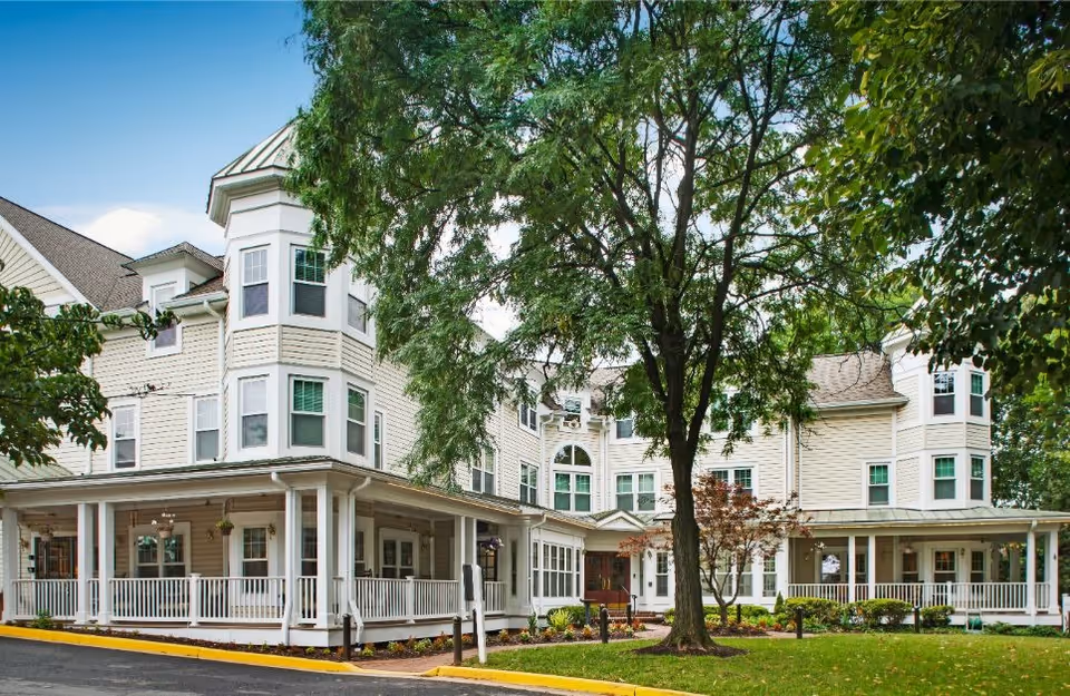 Exterior view of a large, multi-story senior living facility with beige siding and white trim. The building features multiple windows, a wraparound porch with white railings, and is surrounded by trees and landscaped greenery under a clear blue sky.