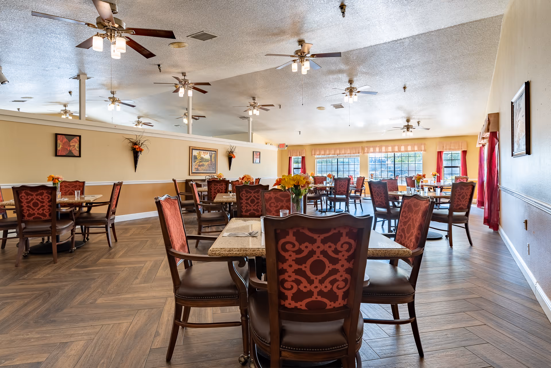 A spacious dining room with multiple tables and chairs arranged neatly. The chairs have red patterned upholstery and the tables are set with glasses and napkins. The room features ceiling fans with lights, large windows with red curtains allowing natural light to fill the space, and wall decorations including paintings and floral arrangements.
