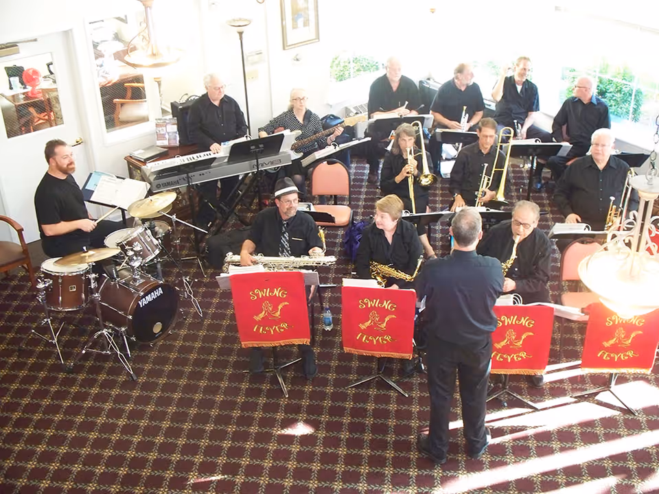 A musical band performing indoors with various musicians playing instruments including drums, keyboard, saxophones, trombones, and trumpets. The band members are seated on chairs arranged in rows, with a conductor standing in front of them. The room has patterned carpet flooring and large windows letting in natural light.