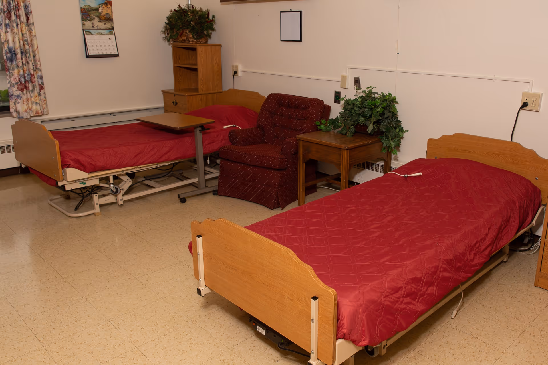 A room with two hospital-style beds covered with red bedspreads, a maroon armchair between the beds, a wooden side table with a green plant, a wooden cabinet with a plant on top, a calendar on the wall, and floral curtains on the window.