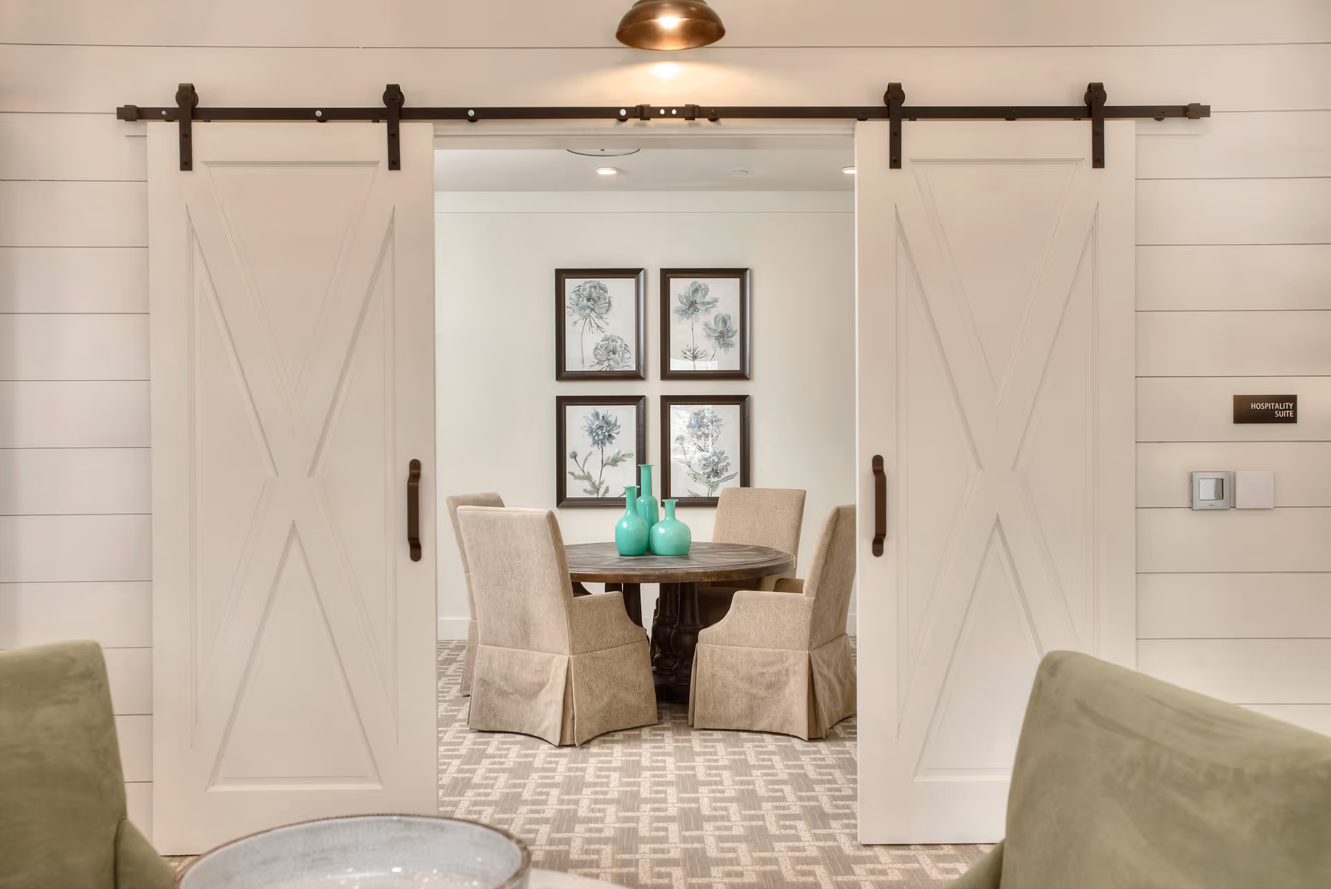A cozy interior room with two white sliding barn doors open to reveal a round wooden table surrounded by four beige upholstered chairs. On the table are three turquoise vases. The back wall features four framed botanical prints arranged in a grid. The room has a patterned carpet and soft lighting.
