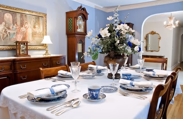 A formal dining room with a table set for six people. The table is covered with a white tablecloth and set with blue and white china, crystal glasses, silverware, and white napkins with blue napkin rings. A large floral centerpiece with white and blue flowers is in the middle of the table. In the background, there is a wooden sideboard with a lamp and a clock, a grandfather clock, a framed painting on the wall, and an archway leading to another room with a chandelier and a mirror.