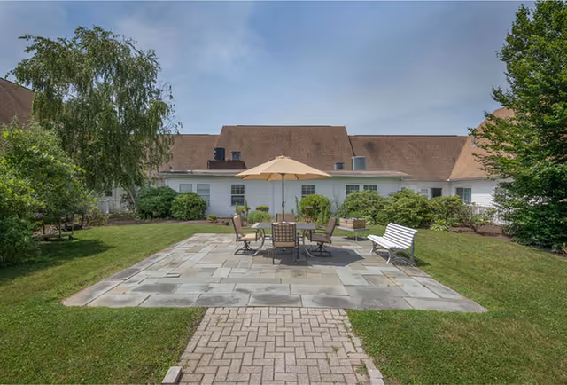 Outdoor patio area with a stone tile floor, a table with an umbrella, four chairs, and a white bench surrounded by green grass, bushes, and trees with a building in the background under a blue sky.