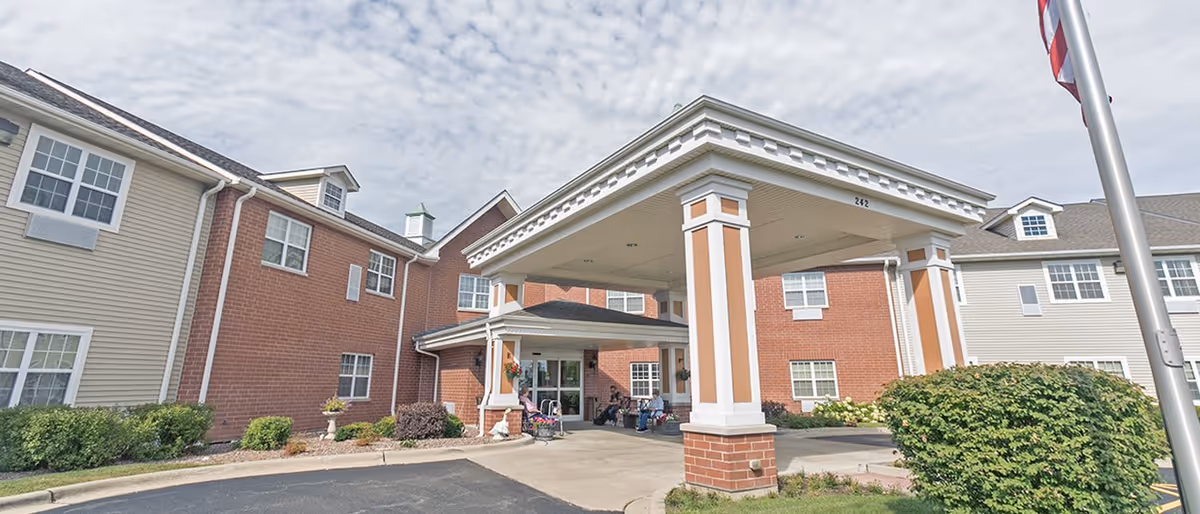 Front entrance of a brick-and-siding senior living building with a covered porte-cochere and flagpole.