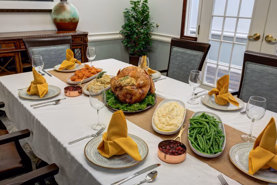 A dining table set for six with a white tablecloth and yellow folded napkins on plates. The table features a roasted turkey centerpiece on a bed of lettuce, mashed potatoes, green beans, cranberry sauce, stuffing, and sweet potatoes. The room has wooden chairs with gray cushions, a wooden sideboard with a decorative vase, a potted plant in the corner, and French doors in the background.