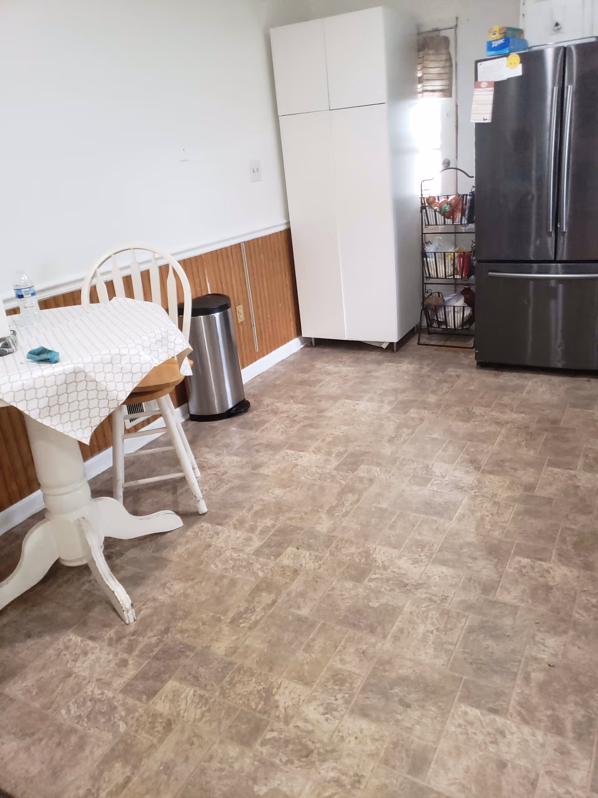 Small kitchen/dining area with a round pedestal table and chair, stainless steel refrigerator, tall white cabinet, trash can, and vinyl tile floor.