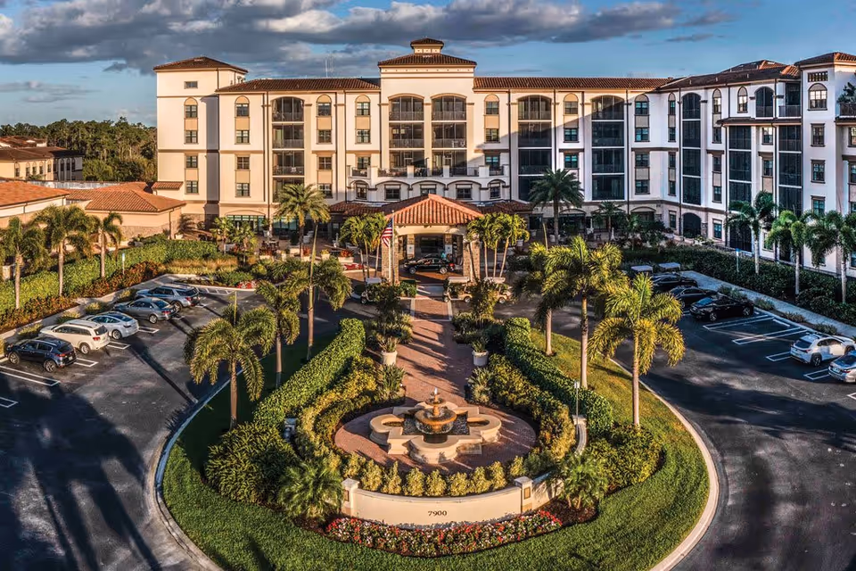 Front exterior view of The Arlington of Naples senior living facility with a circular driveway, landscaped garden featuring a central fountain, palm trees, and parked cars under a partly cloudy sky.