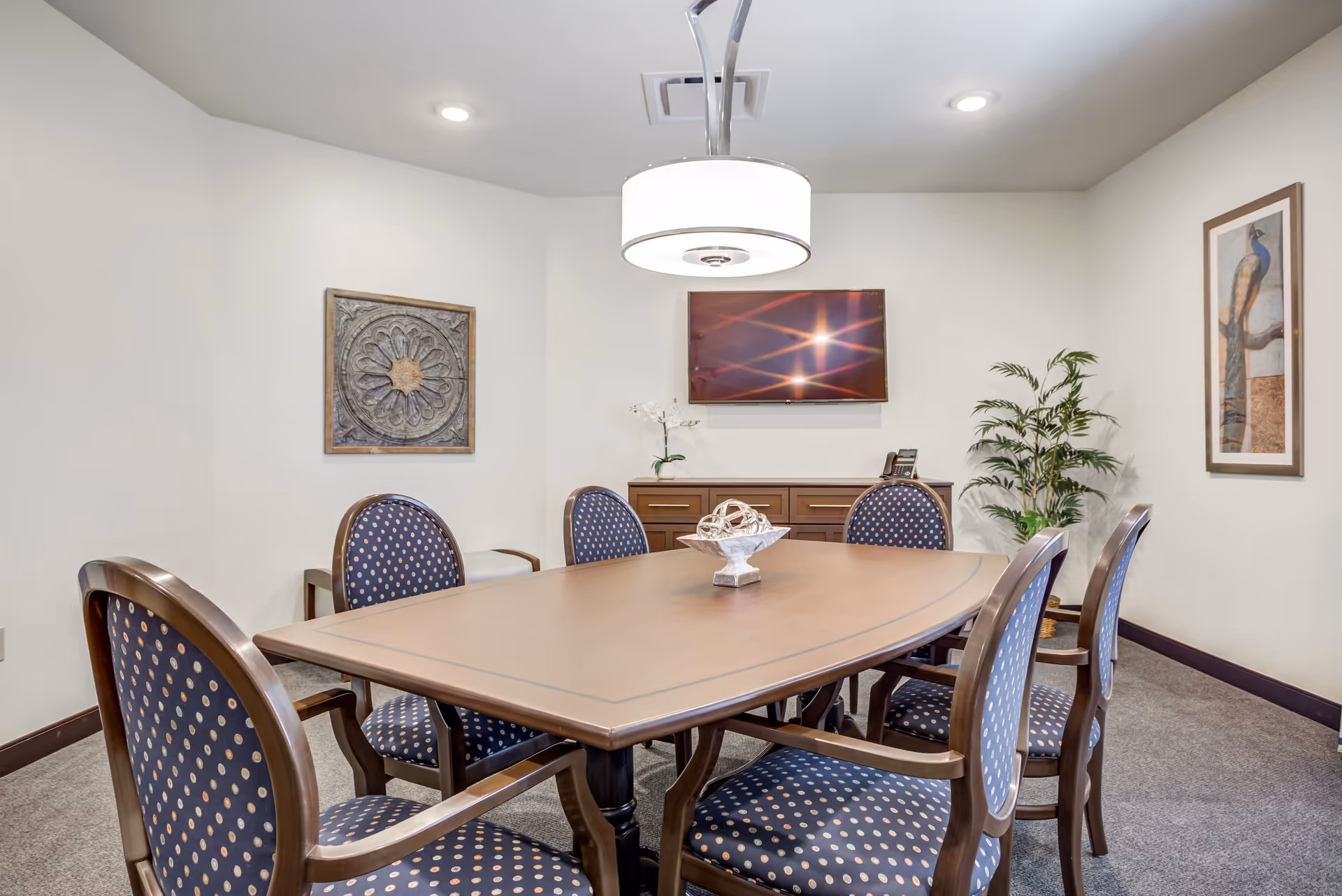 A well-lit meeting room with a large wooden table surrounded by upholstered chairs, a wall-mounted TV, and decorative artwork and plants.