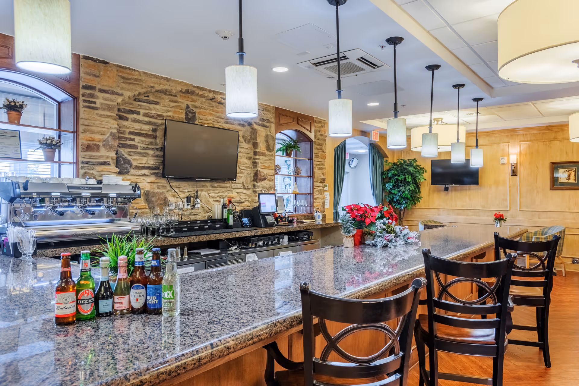 Interior view of a bar area in a senior living facility with a granite countertop, several bottles of beer lined up, three wooden bar stools, hanging pendant lights, a stone accent wall with a mounted flat-screen TV, and shelves with decorative plants and glassware.