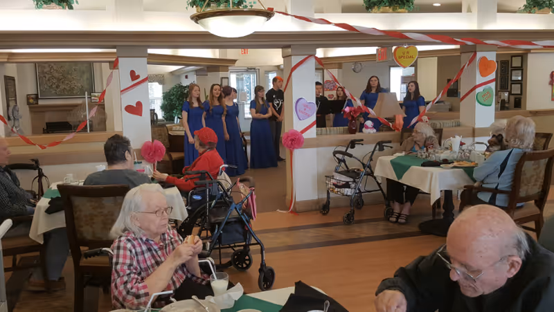 Senior living residents seated at decorated dining tables watching a small group of women in blue dresses perform.