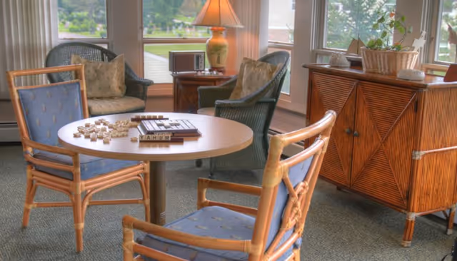 A cozy sitting area with a round wooden table and four wicker chairs with blue cushions. On the table, there is a Scrabble game set up. Behind the table, there are two wicker armchairs with cushions, a wooden cabinet with decorative items, and large windows letting in natural light.