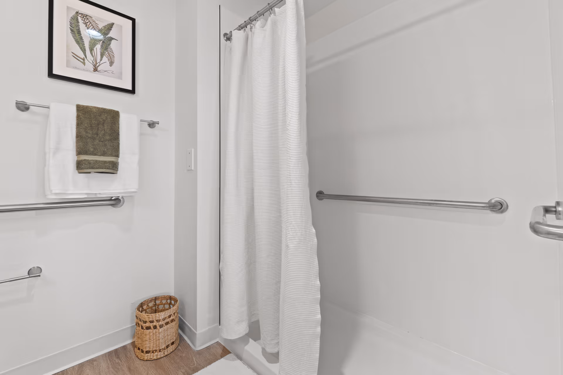 Bathroom showing a shower/tub with a white curtain and grab bars, a towel rack with folded towels and framed artwork, and a wicker wastebasket.