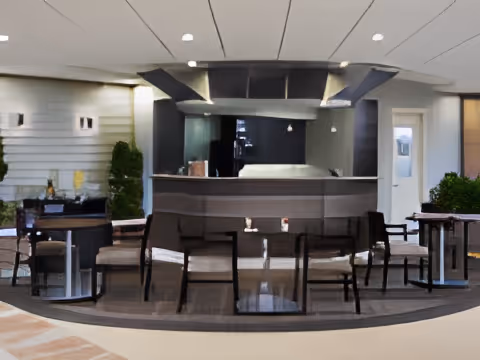 Interior view of a modern common area with a curved bar or counter in the center, surrounded by several chairs and small tables. The space has a clean, contemporary design with neutral colors, plants on either side, and recessed lighting in the ceiling.