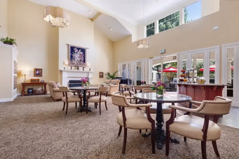 A spacious and well-lit common area with beige walls and carpeted floor, featuring several round tables with cushioned chairs arranged around them. There is a fireplace with a floral tapestry above it, a side table with a lamp and framed pictures, and large windows and glass doors letting in natural light. Outside, red umbrellas are visible on a patio area.