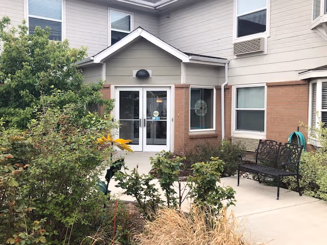 Front entrance of a senior living building with double glass doors, a small landscaped courtyard and a bench.