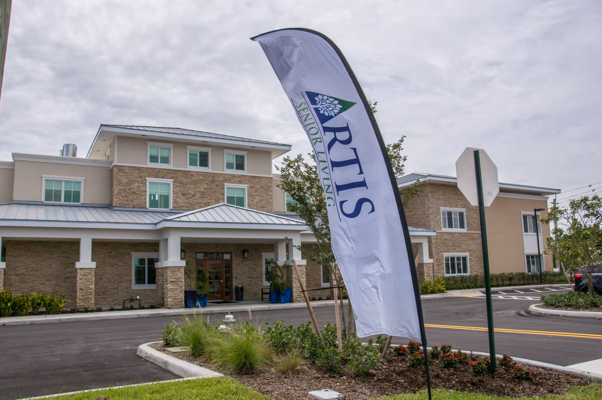 Exterior view of a modern senior living facility building with beige and stone facade, multiple windows, and a metal roof. In front of the building, there is a tall white flag with the text 'ARTIS SENIOR LIVING'. The area includes a paved road, landscaped greenery, and a stop sign.