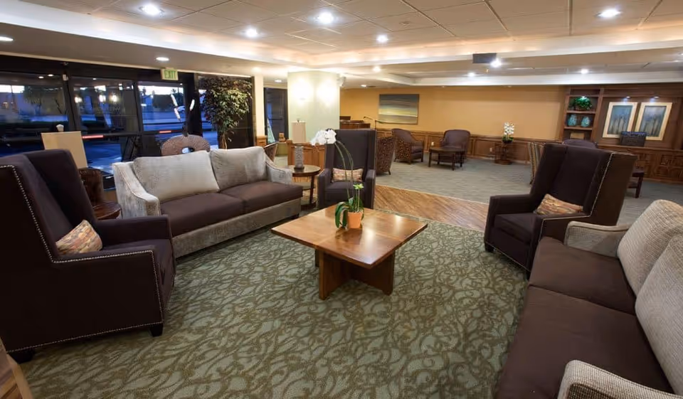 A spacious and well-lit living room area in a senior living facility featuring multiple cushioned sofas and armchairs arranged around a wooden coffee table with a potted orchid plant. The room has patterned carpet flooring, wood paneling on the walls, framed artwork, and large windows letting in natural light.