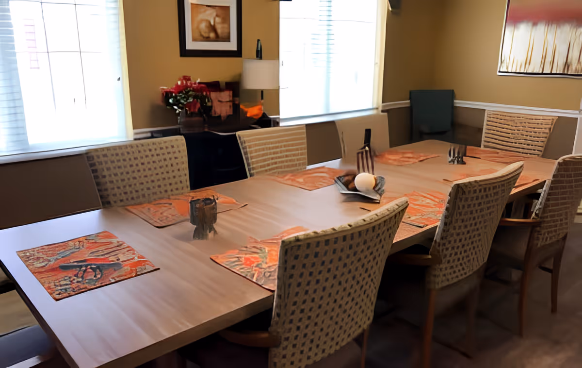 A dining room with a long rectangular wooden table set with orange patterned placemats and decorative items. The room has several cushioned chairs with patterned upholstery around the table. There are two windows with white blinds letting in natural light, a sideboard with a lamp and flowers, and framed artwork on the walls.