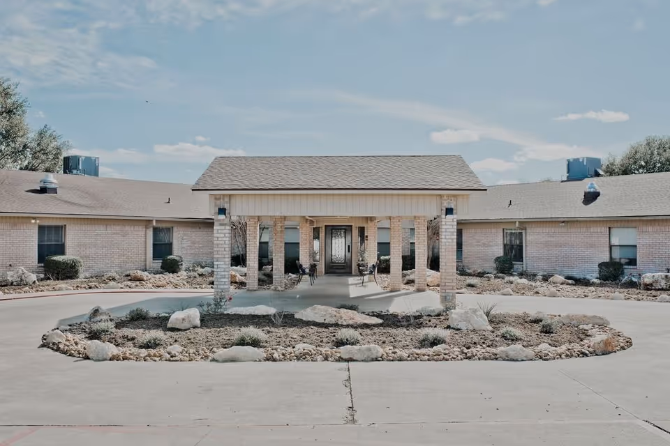 Front exterior view of Hallettsville Nursing and Rehabilitation Center showing a single-story brick building with a covered entrance supported by brick pillars, surrounded by a circular driveway and landscaped with rocks and small shrubs under a partly cloudy sky.