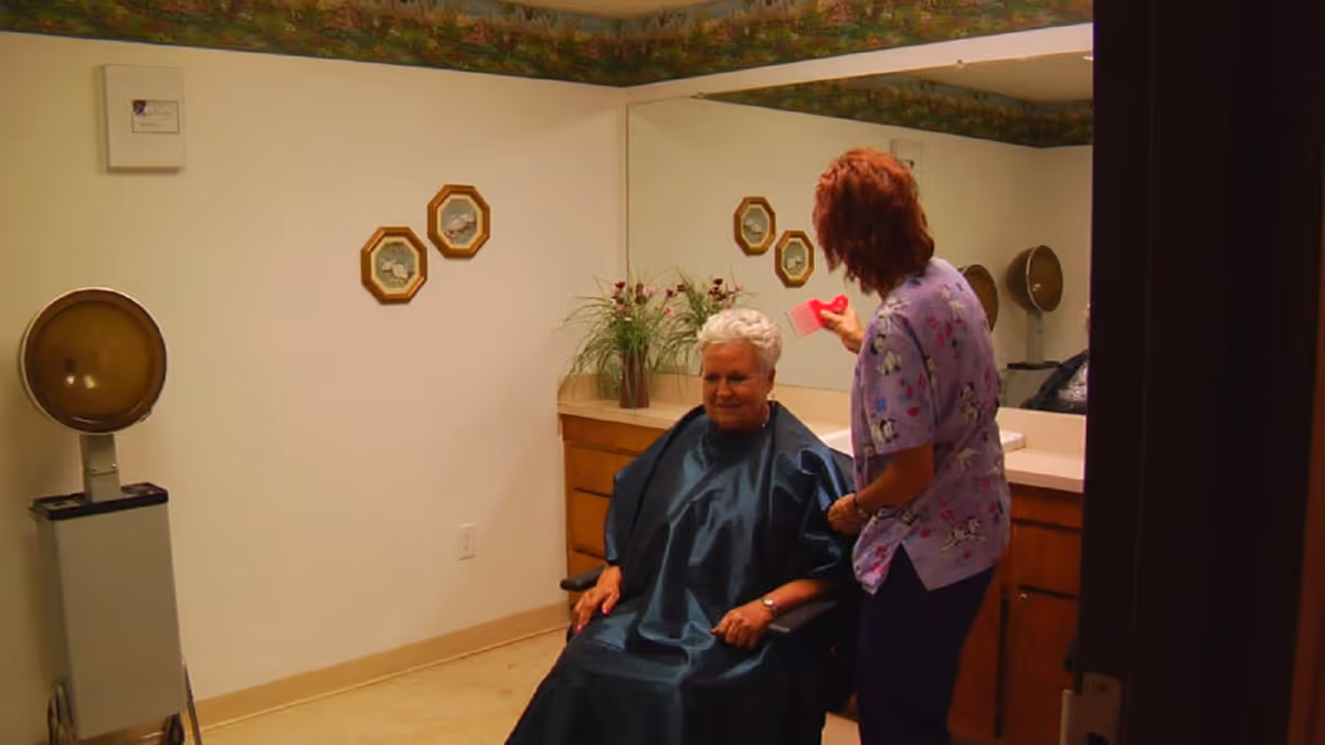 An elderly woman sitting in a chair with a hairdressing cape on while a caregiver or stylist stands beside her holding a comb in a room with a large mirror, wooden cabinets, and a vintage hair dryer.