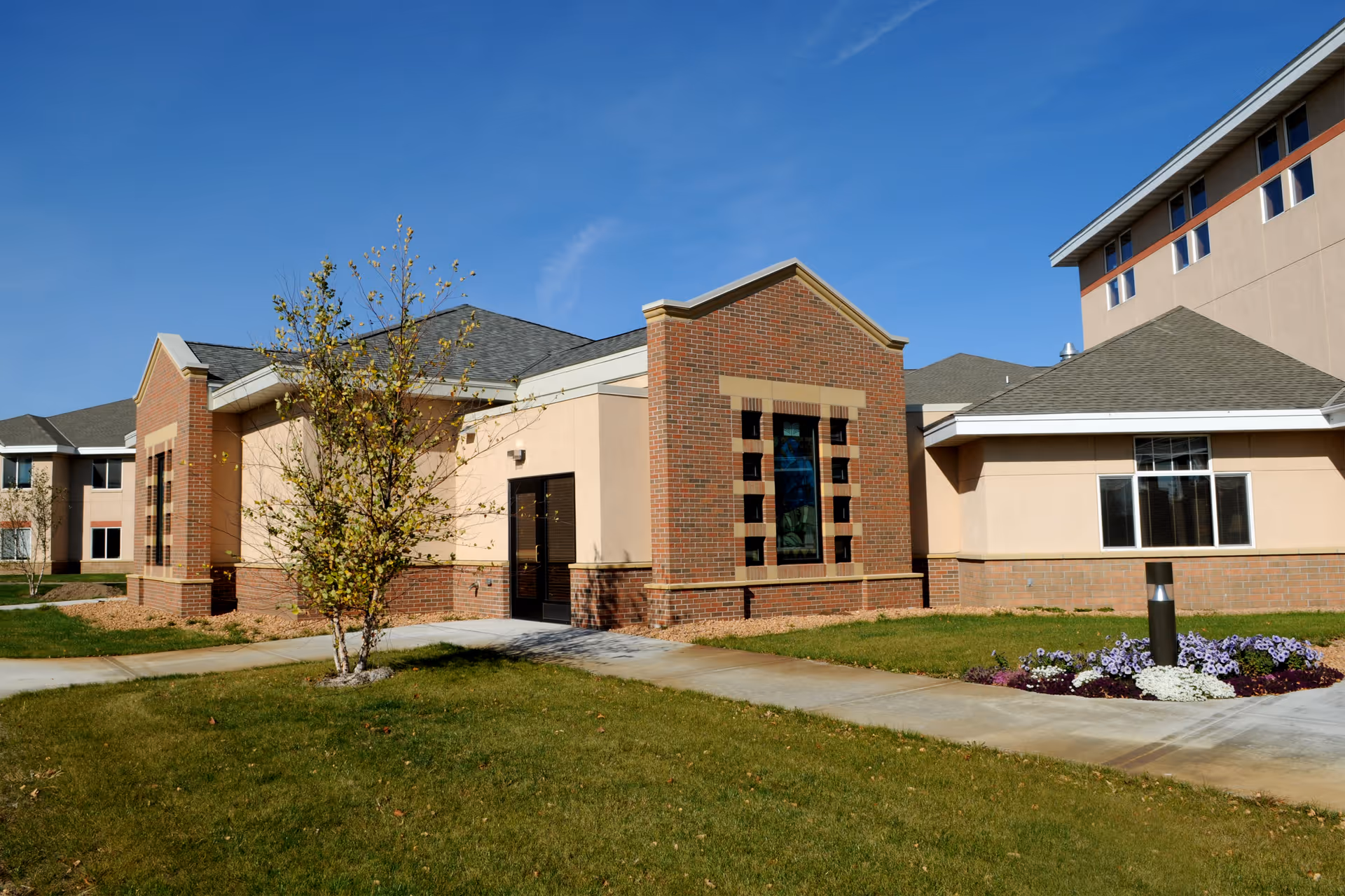 Exterior view of a senior living facility building with beige and brick walls, multiple windows, a small tree, a flower bed, and a paved walkway under a clear blue sky.