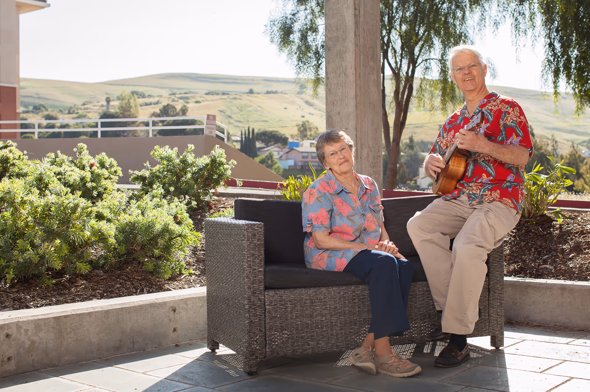 An elderly woman sitting on a wicker outdoor sofa and an elderly man standing next to her playing a ukulele. They are outside on a patio with greenery and hills in the background, enjoying a sunny day.