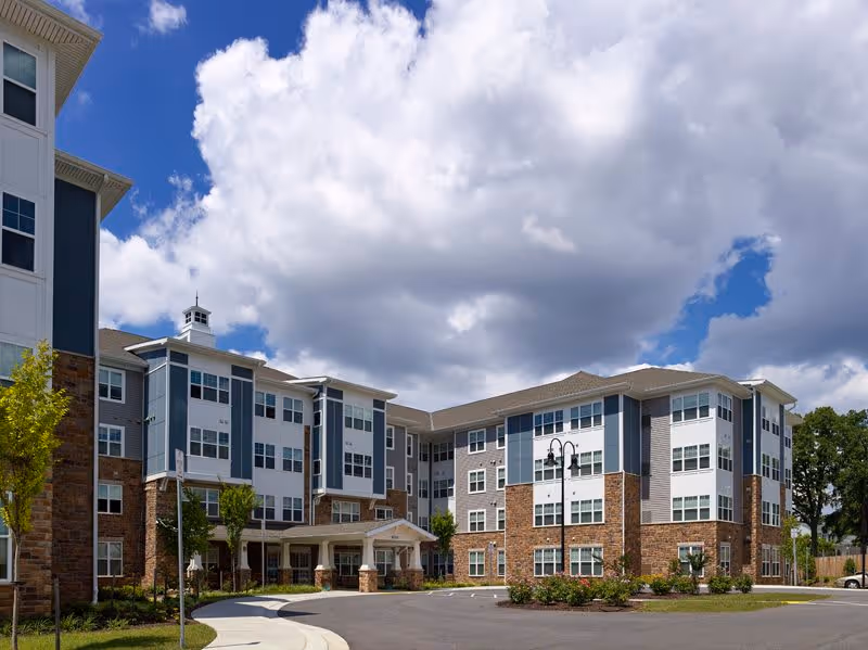 Exterior view of a multi-story senior living facility building with a mix of brick and siding facade under a partly cloudy blue sky. The building has many windows and a covered entrance with a driveway and landscaped greenery around.